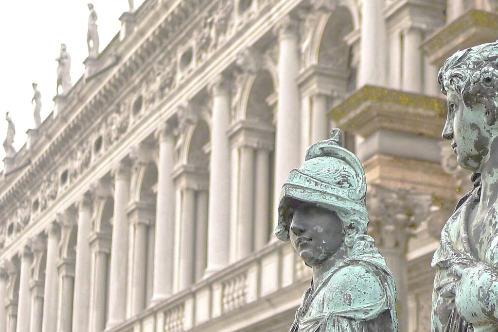 Das ist Bild 1 von 3 - Im Vordergrund stehen zwei historische Bronzestatuen mit grünlicher Patina, deren Oberfläche durch Zeit und Witterung geprägt ist. Eine der Figuren trägt einen Helm und wirkt wie ein Wächter, während daneben eine zweite Figur ruhig in die Ferne blickt. Hinter den Statuen erhebt sich eine elegante Fassade mit zahlreichen Bögen, Säulen und dekorativen Details – typische Renaissance-Architektur rund um den Markusplatz im Herzen von Venedig. Diese Architektur erinnert an die Zeit der mächtigen venezianischen Republik, als die Stadt ein bedeutendes Handelszentrum zwischen Europa und dem Orient war. Für Besucherinnen und Besucher aus aller Welt liegt genau darin ein großer Teil der Faszination: Venedig verbindet Geschichte, Kunst und einzigartige Architektur mit der besonderen Atmosphäre einer Stadt auf dem Wasser. Die Kombination aus Kanälen, Palästen und lebendiger Geschichte macht den Ort für viele Menschen zu einem der schönsten Reiseziele der Welt. Der nachfolgende sichtbare Text ist der gleiche wie in der barrierefreien Bildbeschreibung.